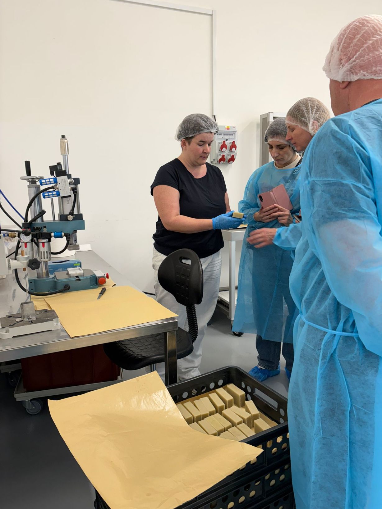 Na pracovním stole jsou rozložené kartonové krabičky NATAVA s průhledným okénkem, do kterých jsou ručně vkládány olejové koupelové koule s bylinným povrchem. Vedle nich leží role etiket a další připravené produkty. Prostor působí jako čistá balicí nebo dokončovací zóna menší kosmetické výroby.