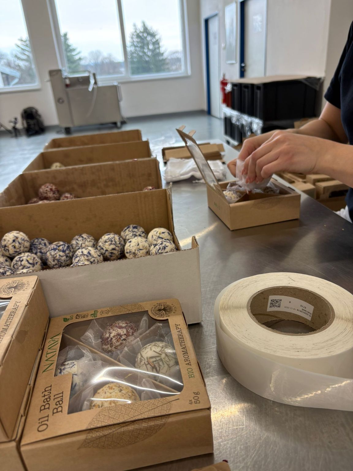Pracovník ručně vkládá bylinné olejové koule do přírodně laděných krabiček NATAVA. Na stole jsou připravené další obaly, etikety a zásobníky s produkty. Místnost s velkými okny a modrými dveřmi působí jako moderní, světlý provoz určený pro finální balení přírodní kosmetiky.