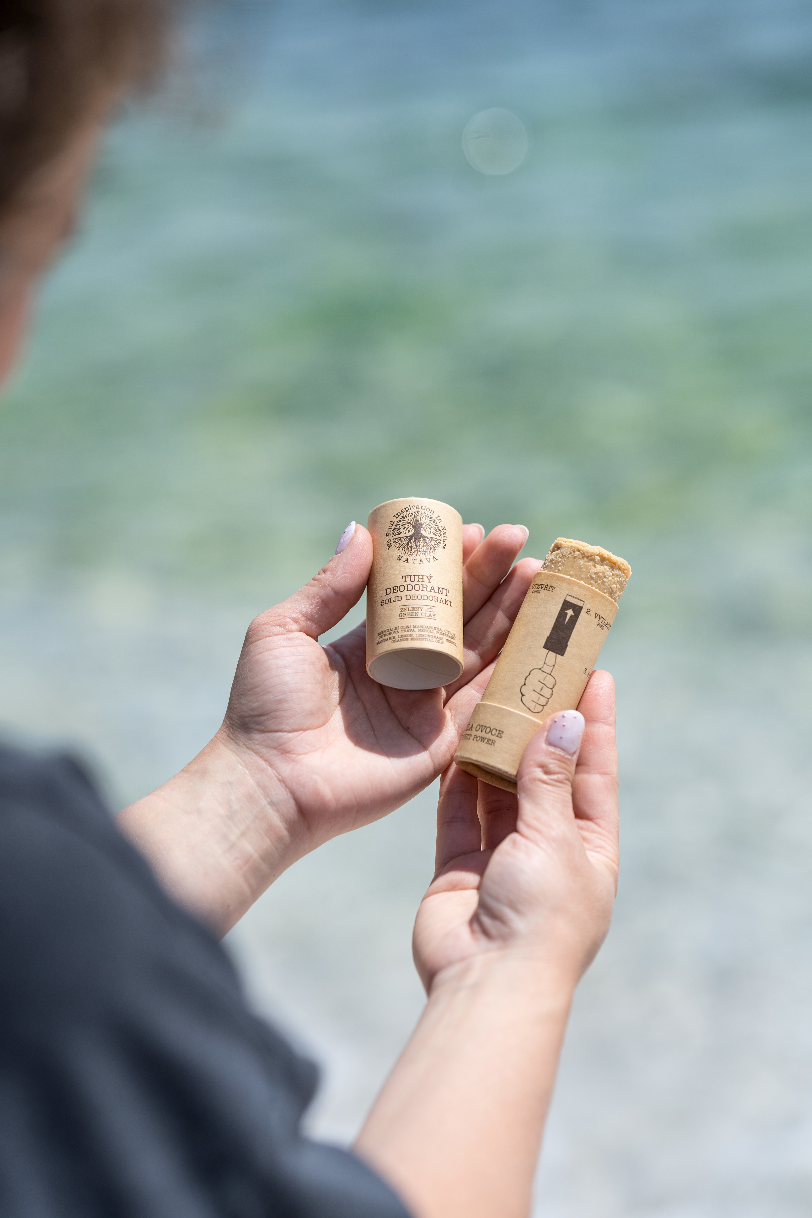 Tuhé deodoranty NATAVA v papírovém tubusu držené v ruce u vody jako příklad travel friendly kosmetiky